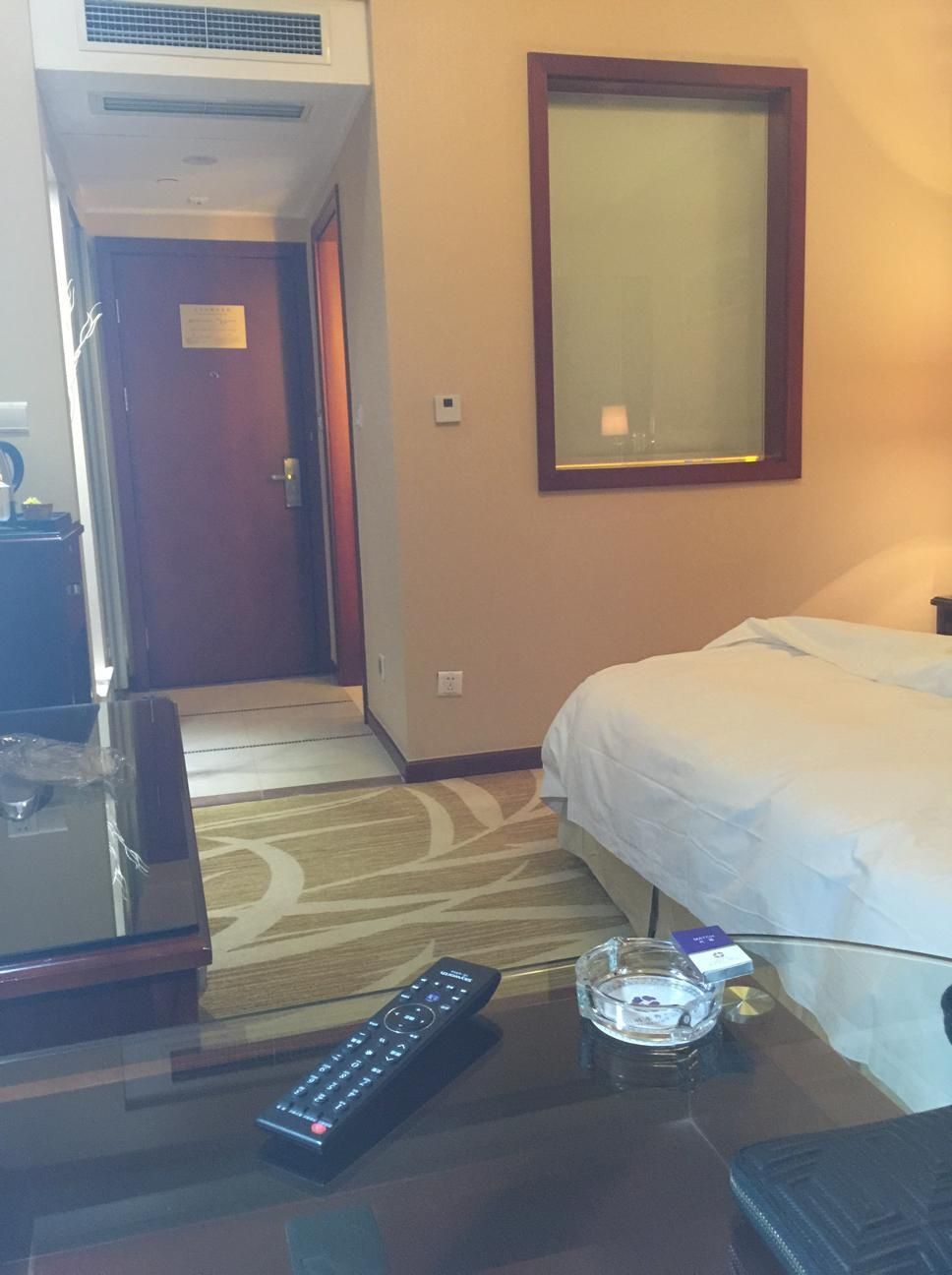 A hotel room with a bed, a desk with a remote control, a glass ashtray, and a door leading to another room.