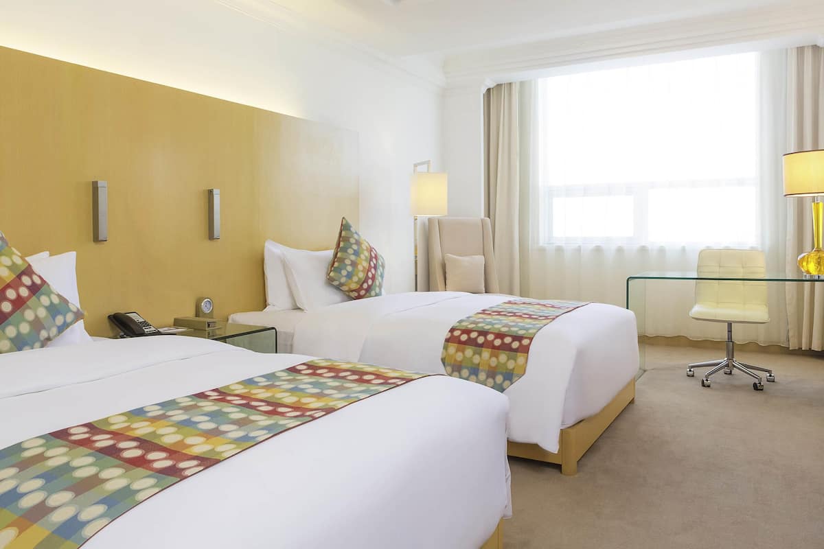 A hotel room with two beds, a desk, a chair, a lamp, and a window with curtains.