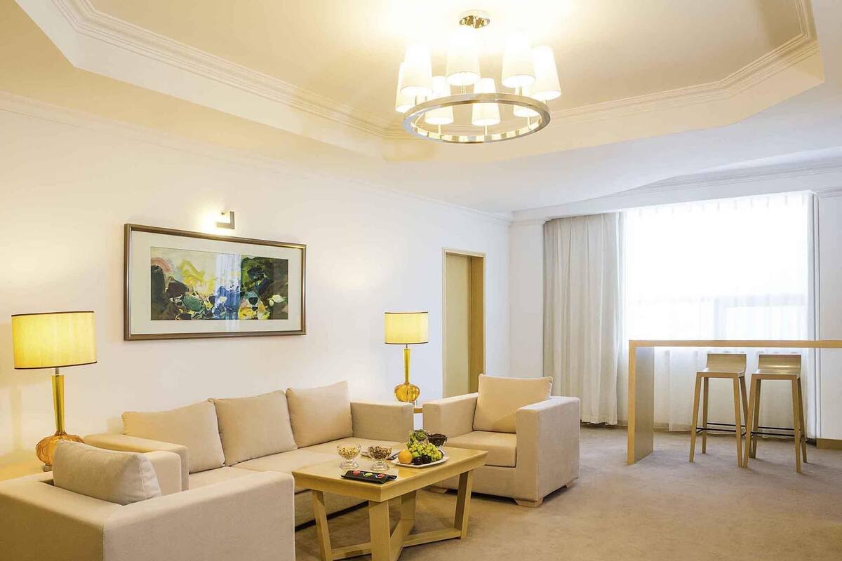 A hotel room with a beige sofa set, a wooden coffee table with fruits, two lamps, a painting on the wall, and a view of the city through the window.