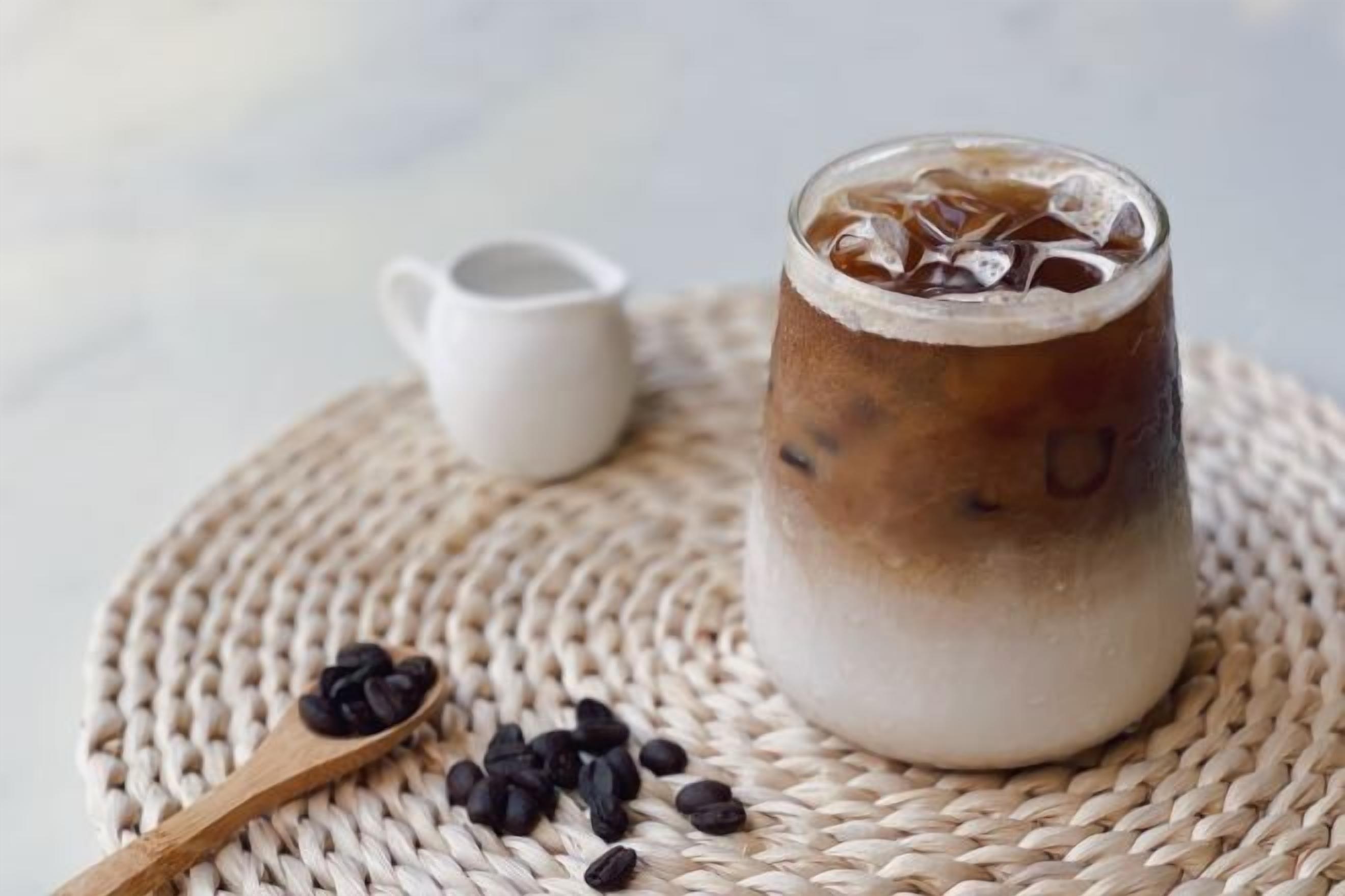 Un vaso de café helado con cubitos de hielo sobre un posavasos tejido, acompañado de una cuchara de madera con granos de café y una taza blanca pequeña.