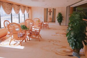 Habitación de hotel con zona de comedor que incluye sillas de mimbre y una mesa, una planta grande en maceta y suelo alfombrado.