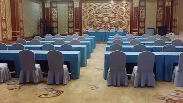 Een congreszaal met rijen tafels en stoelen, een podium met een spreker en decoratieve wandversieringen.