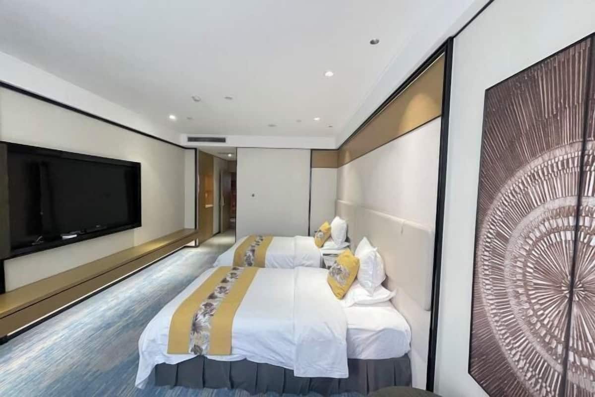 A hotel room with a large flat-screen TV mounted on the wall, a bed with white bedding and decorative pillows, and a large abstract artwork on the wall.