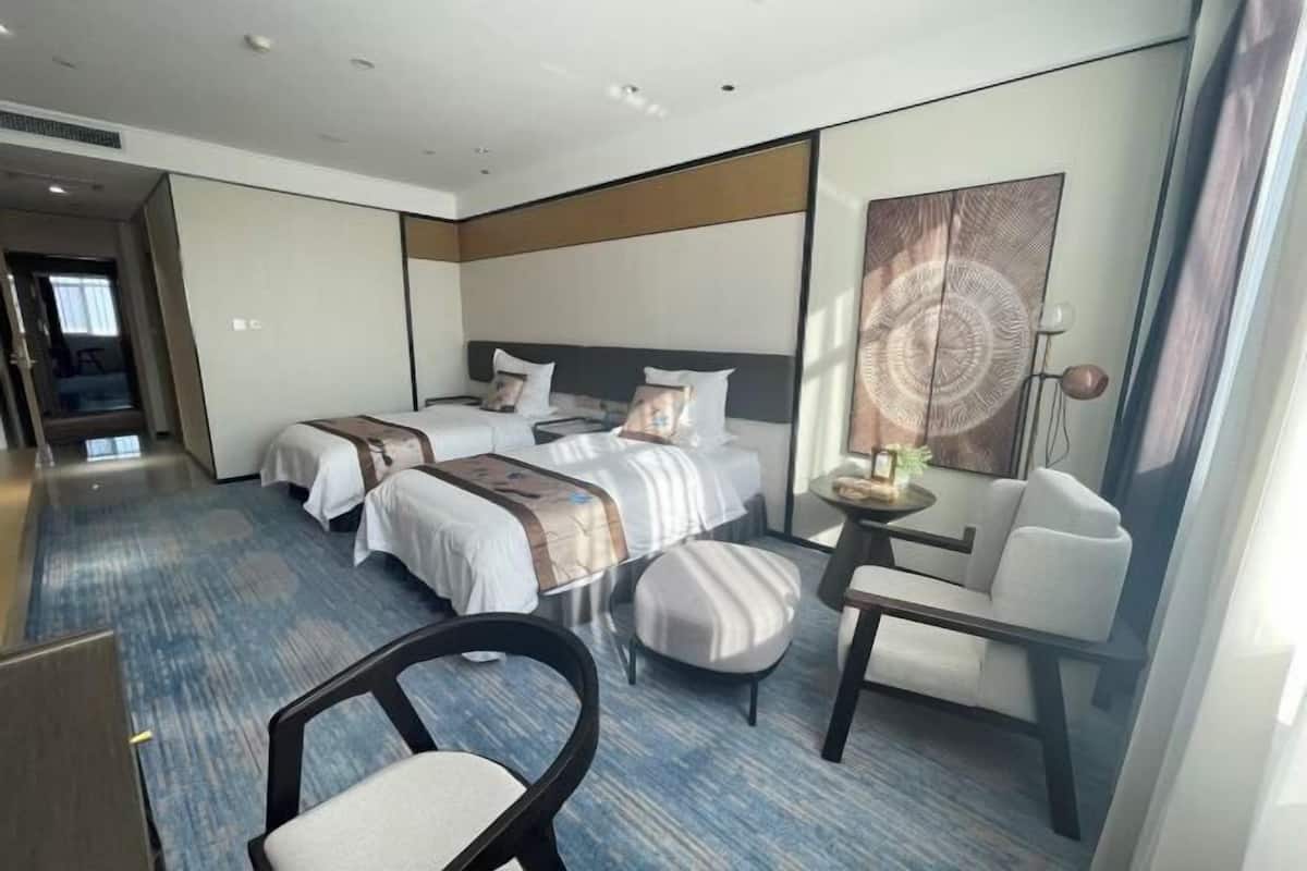A hotel room with two beds, a desk, a chair, and a small table with a lamp.