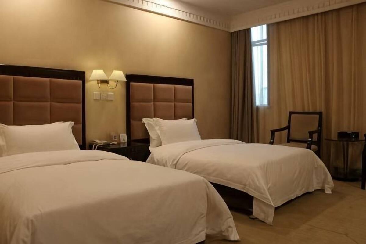 Habitación de hotel con dos camas, una mesita de noche con lámpara, una silla y un ventanal con cortinas.