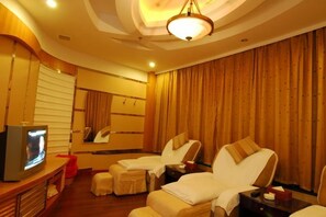Een hotelkamer met een televisie, twee relaxfauteuils, een massagetafel en een groot raam met gordijnen.