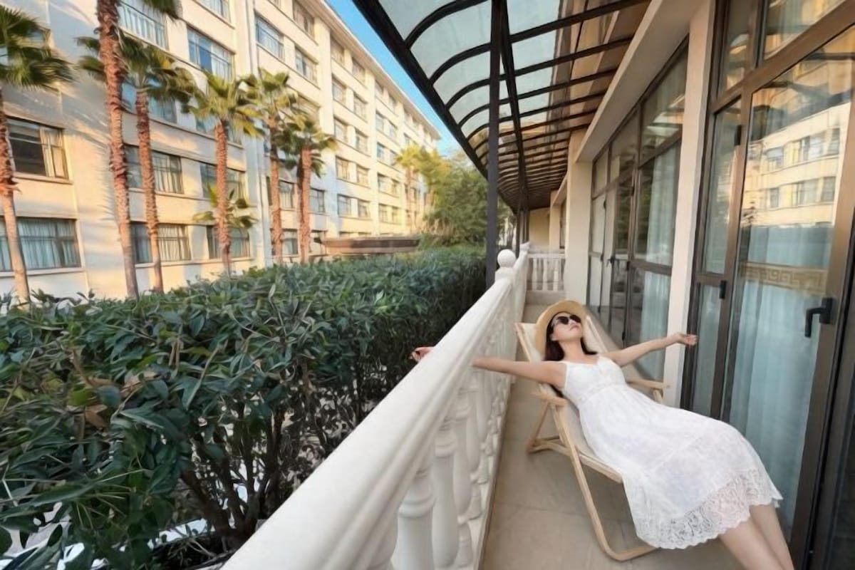 Un balcon avec une balustrade blanche, une personne portant une robe blanche, et un bâtiment avec de nombreuses fenêtres, des palmiers en arrière-plan.
