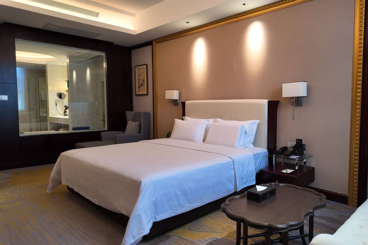 Habitación de hotel con una cama grande, una mesita de noche con un teléfono y una caja, una pequeña mesa redonda, una silla, un espejo y un cuadro enmarcado en la pared.