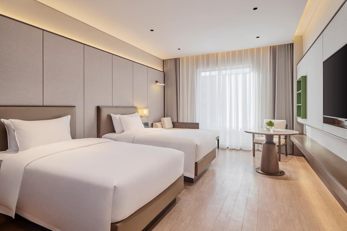 Habitación de hotel con dos camas, una mesa de comedor, televisor y un ventanal con cortinas.