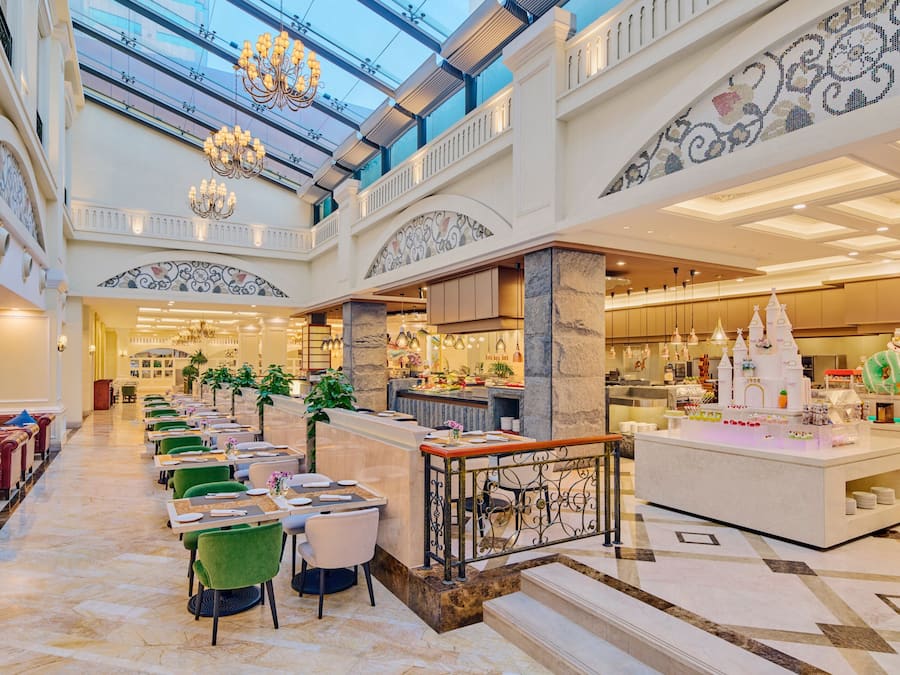 Un vestíbulo de hotel con techo alto, candelabros, una zona de bar central y una escalera que conduce a los pisos superiores.