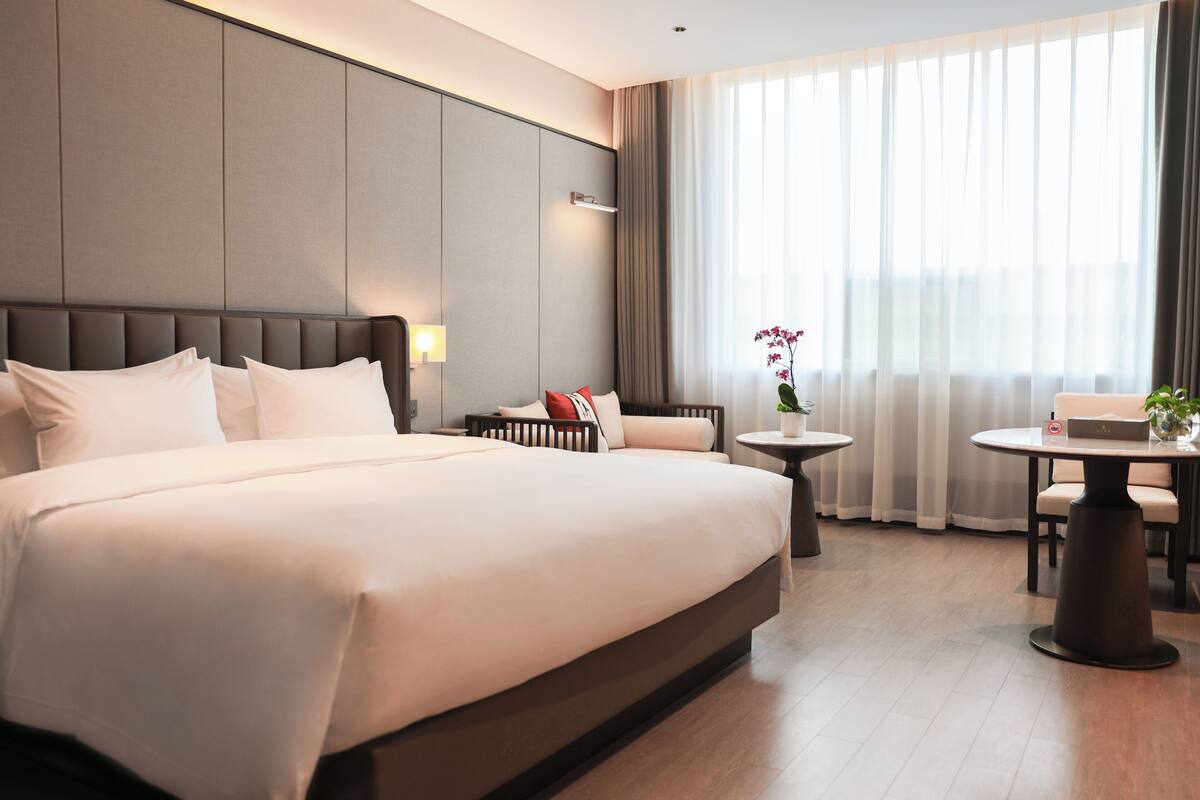 Habitación de hotel moderna con una cama grande, una mesita de noche, una pequeña mesa redonda y vistas al exterior a través de cortinas translúcidas.