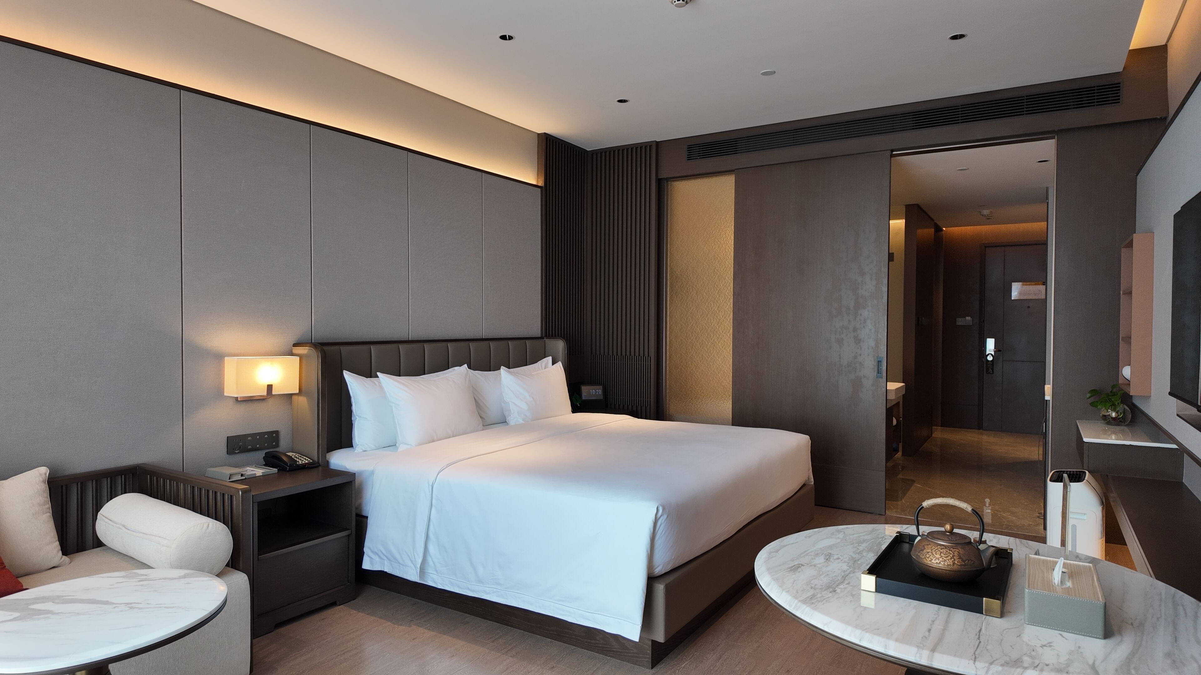 A modern hotel room with a large bed, bedside table, lamp, and a round coffee table.