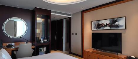 A hotel room with a bed, a desk with a computer, a mirror, a TV, and a minibar.