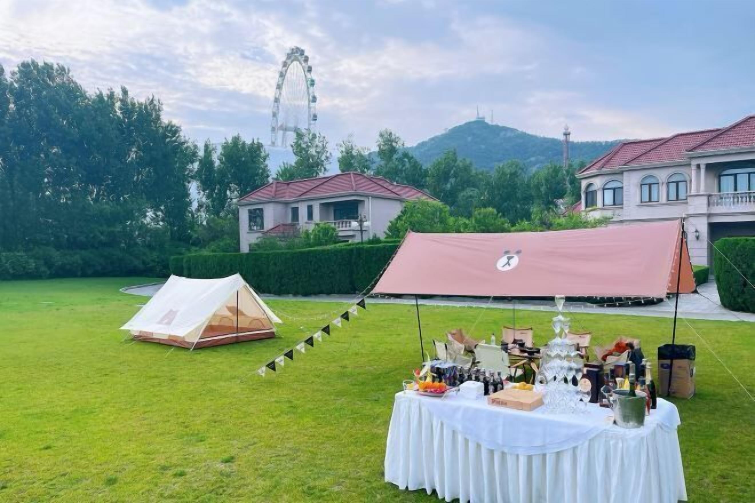 A tented outdoor area with a table set for a meal, surrounded by greenery and buildings in the background.