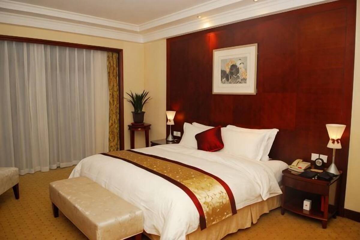A hotel room with a large bed, a bedside table with a lamp, a small table with a plant, and a window with curtains.