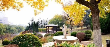 A park with a statue on a circular platform, surrounded by various trees and shrubs.