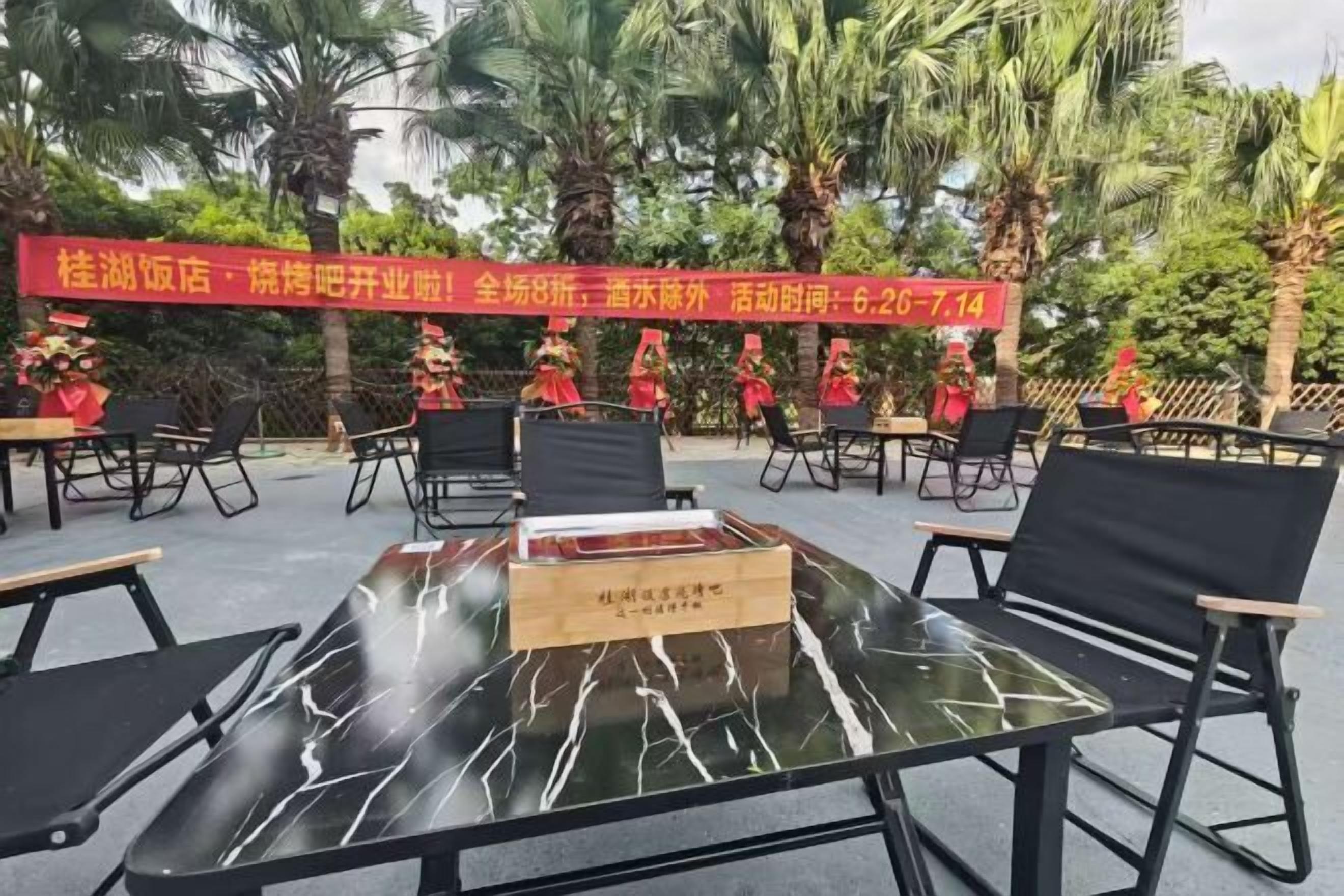 An outdoor seating area with tables and chairs, palm trees in the background, and a red banner with text.