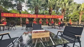 An outdoor seating area with tables and chairs, palm trees in the background, and a red banner with text. - Guilin Park Hotel (GUILIN)