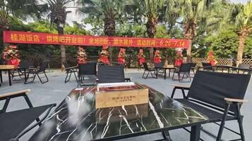 An outdoor seating area with tables and chairs, palm trees in the background, and a red banner with text.