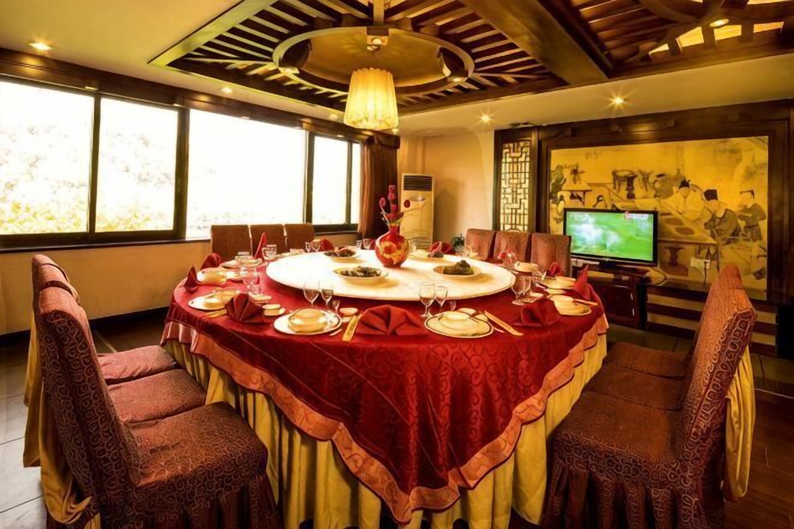 A round dining table with a red tablecloth, surrounded by chairs, in a room with large windows and a television.