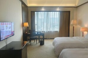 Quarto de hotel com duas camas, uma escrivaninha com cadeira, televisão e uma janela grande com cortinas.