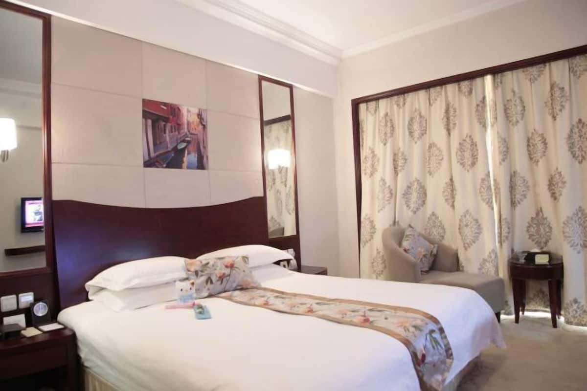 Ein Hotelzimmer mit einem großen Bett, einem Nachttisch, einem Sessel, einem Spiegel und einem Vorhang.