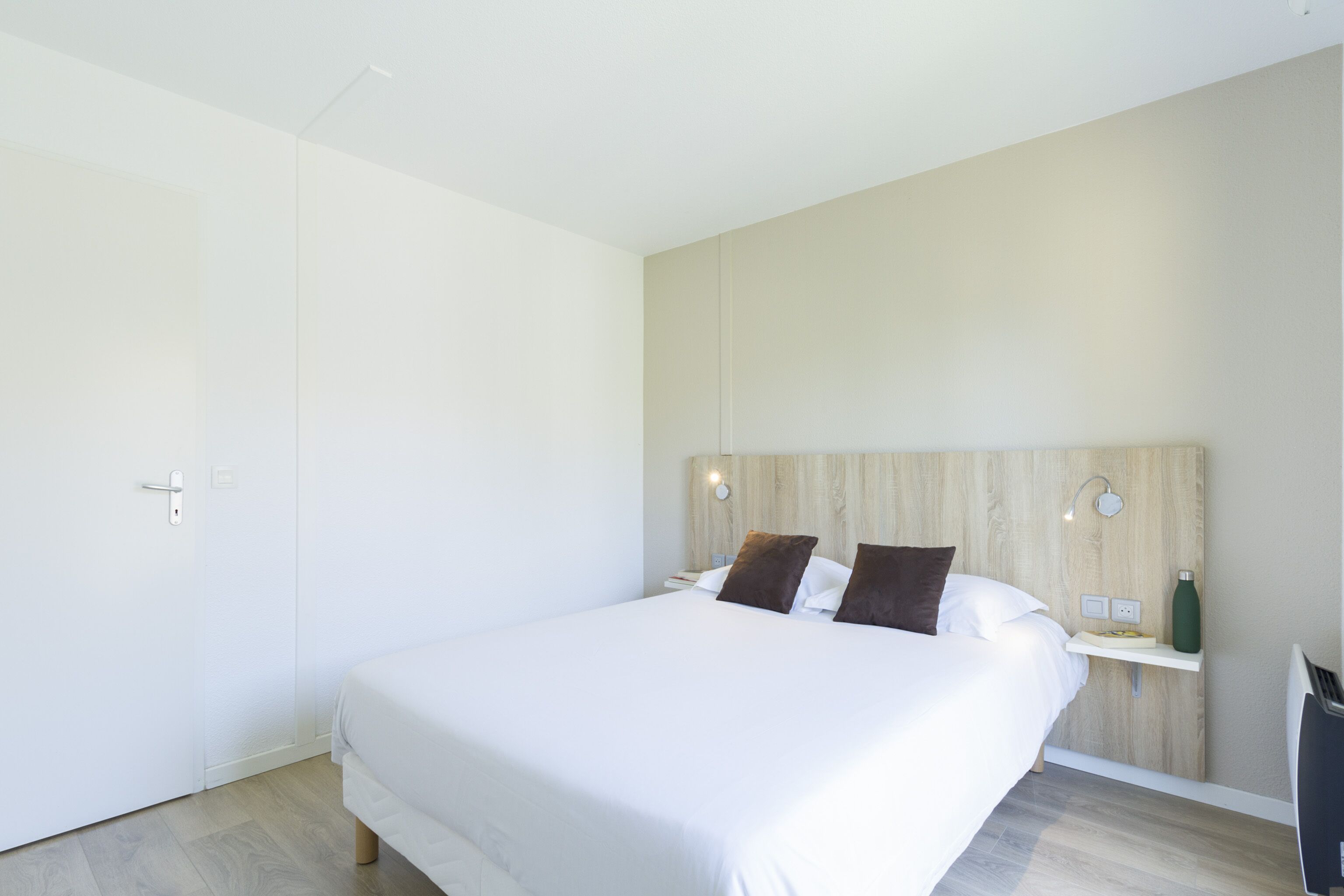 Ett modernt hotellrum med en stor säng, ett träfärgat sänggavel, sänglampor, en liten hylla med en vattenflaska och ett eluttag.