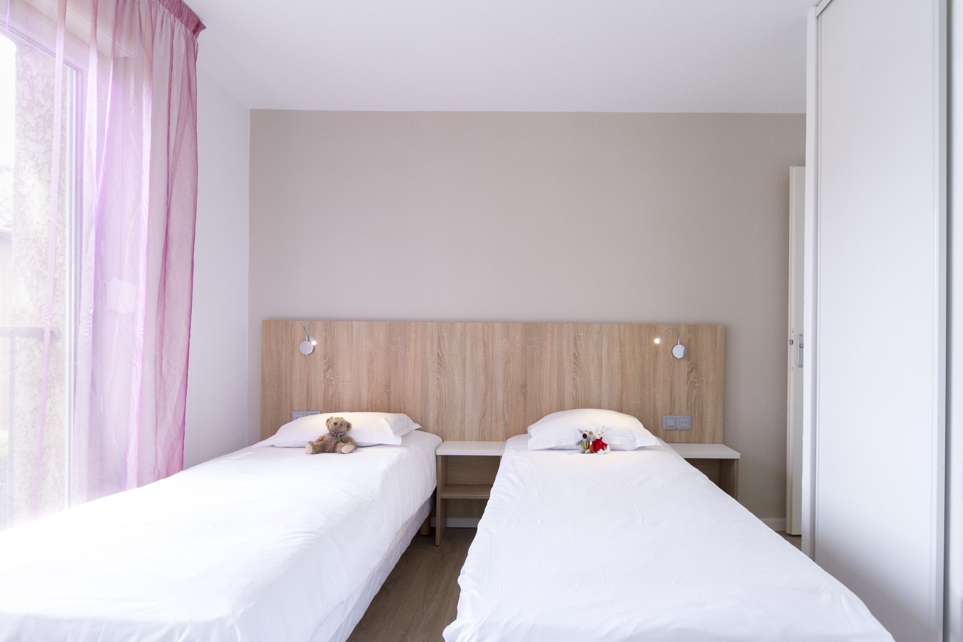 A hotel room with two single beds, a wooden headboard, a nightstand, and a window with pink curtains.
