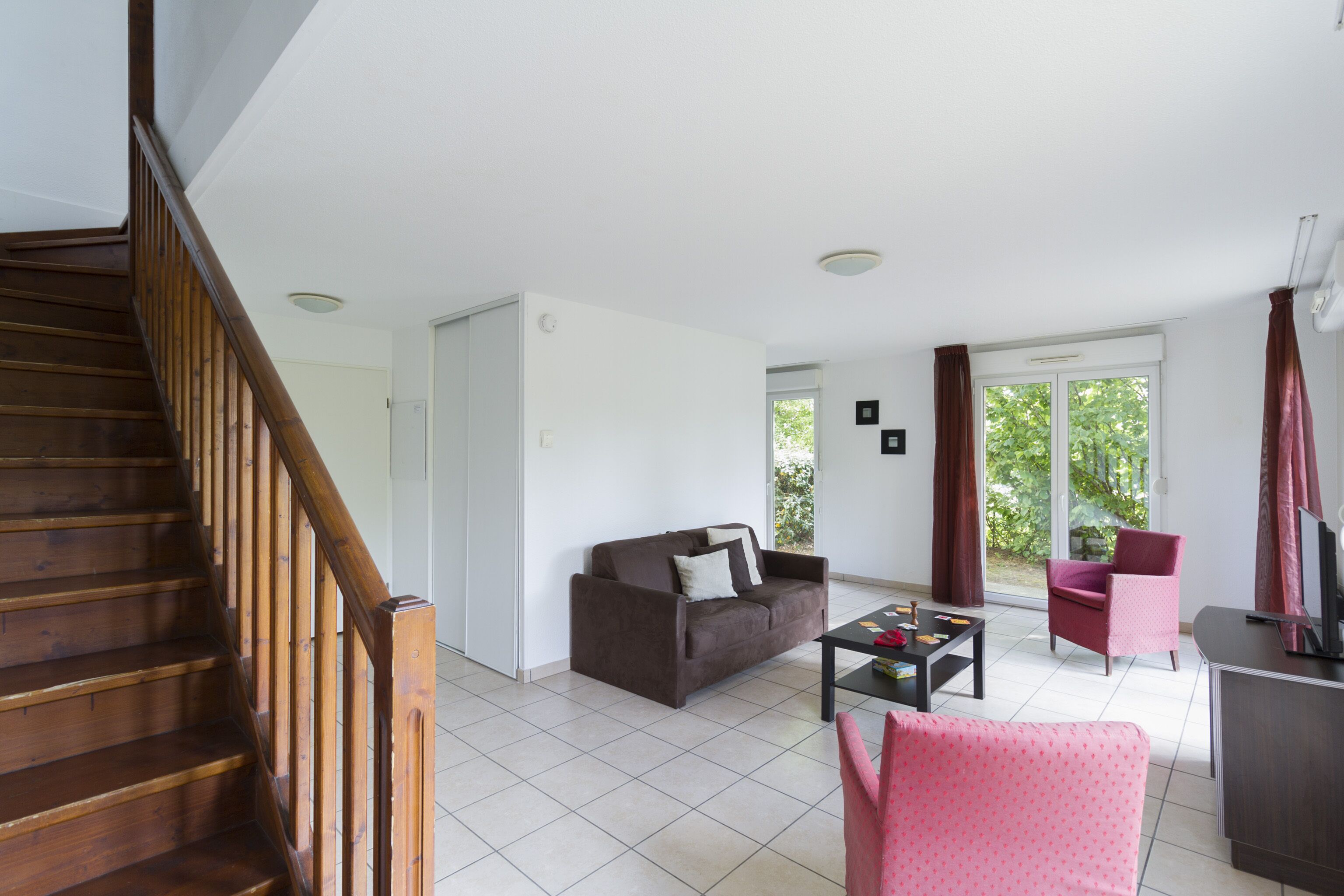 Un salon avec un escalier en bois, un canapé marron, un fauteuil rose et un téléviseur posé sur un meuble.