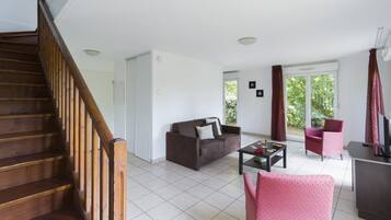 Un salon avec un escalier en bois, un canapé marron, un fauteuil rose et un téléviseur posé sur un meuble.