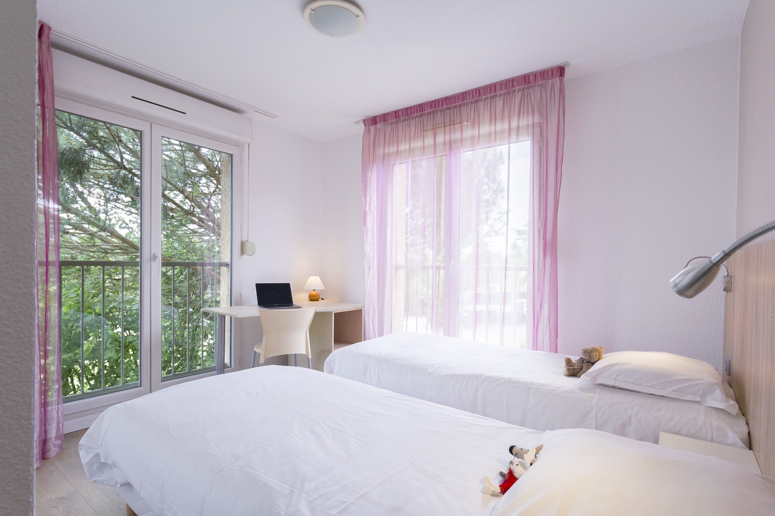 A hotel room with two beds, a desk with a laptop, a lamp, and large windows with pink curtains.