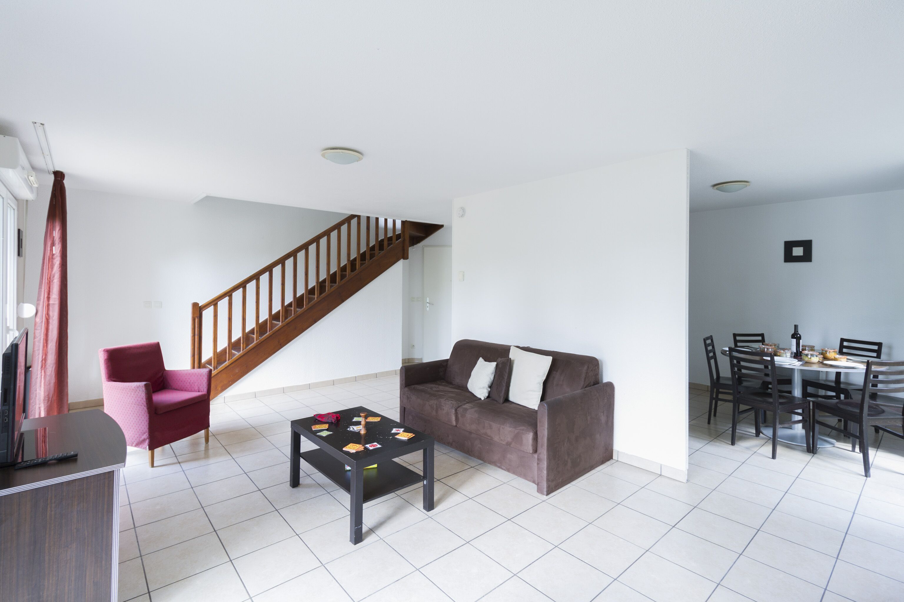 Een ruime woonkamer met een bruine bank, een rode fauteuil, een salontafel en een eettafel met stoelen. Op de achtergrond is een houten trap zichtbaar.