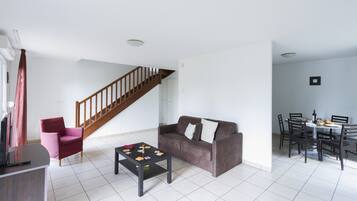 Een ruime woonkamer met een bruine bank, een rode fauteuil, een salontafel en een eettafel met stoelen. Op de achtergrond is een houten trap zichtbaar.