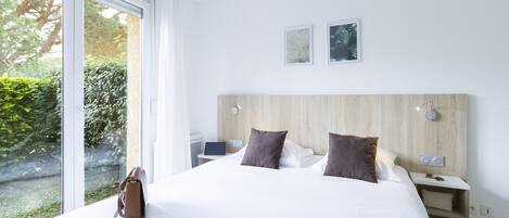Una habitación de hotel moderna con una cama grande, una mesita de noche con lámpara y vistas al exterior a zonas verdes.