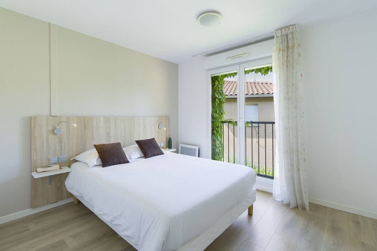 Une chambre à coucher avec un grand lit, des tables de chevet, une commode, une fenêtre avec des rideaux et une vue sur l’extérieur.