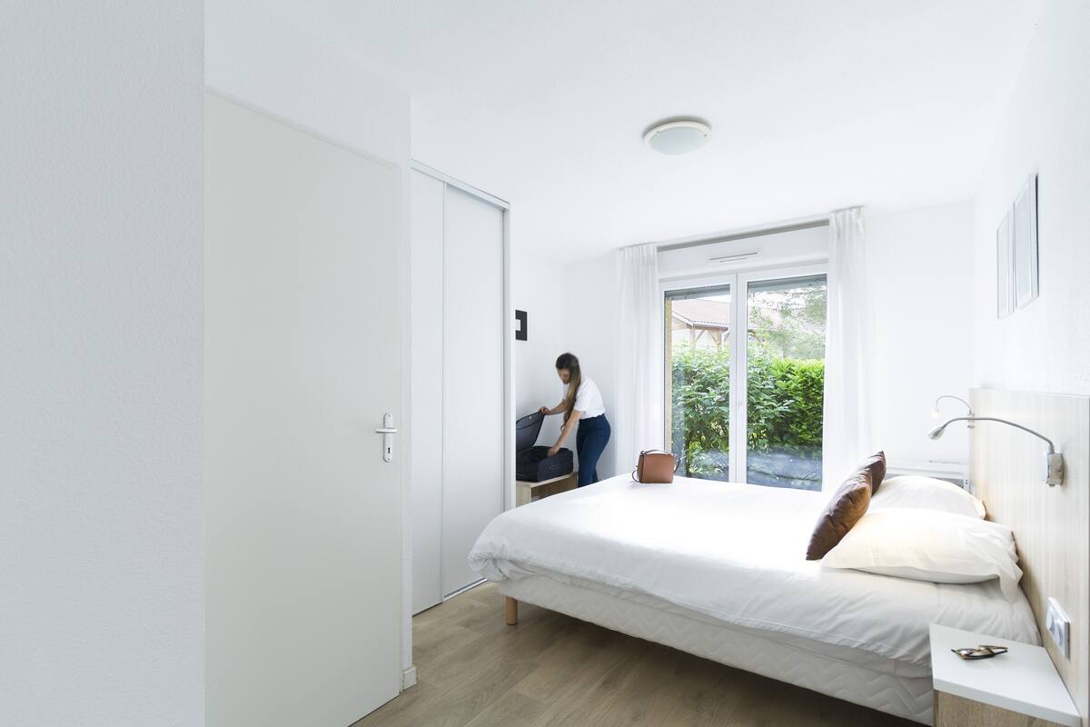 Une chambre à coucher moderne avec un grand lit, une table de chevet, une fenêtre avec des rideaux et une vue sur la verdure à l’extérieur.