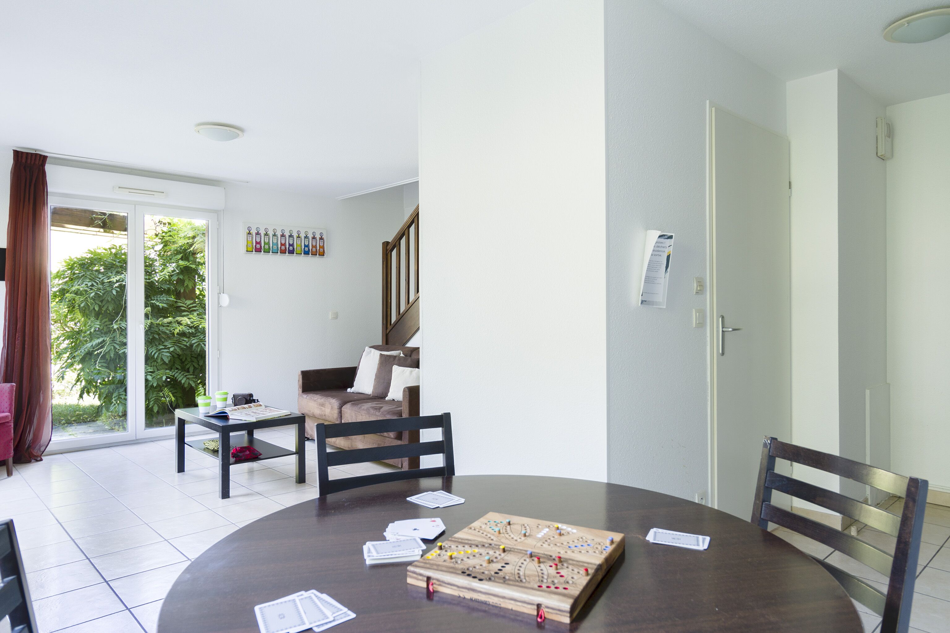 A dining area with a round table, chairs, and a board game. There is a sofa and a staircase in the background.