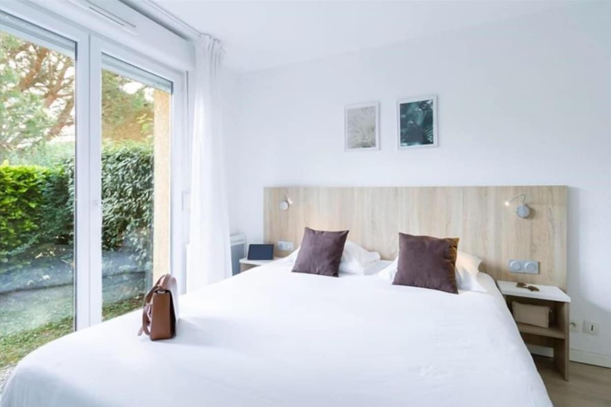 Une chambre d’hôtel moderne avec un grand lit, une tête de lit en bois, deux tables de chevet et une vue sur la verdure.