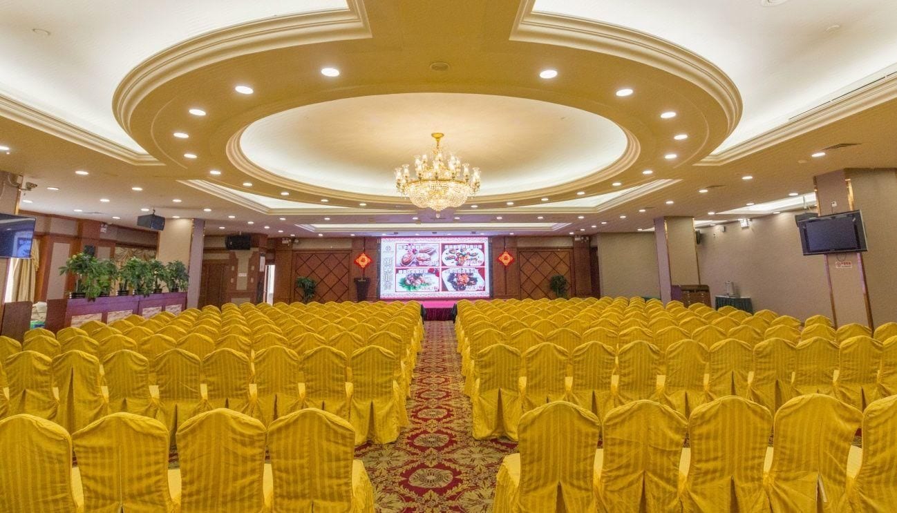 A conference room with rows of yellow chairs, a large screen at the front, and a chandelier.