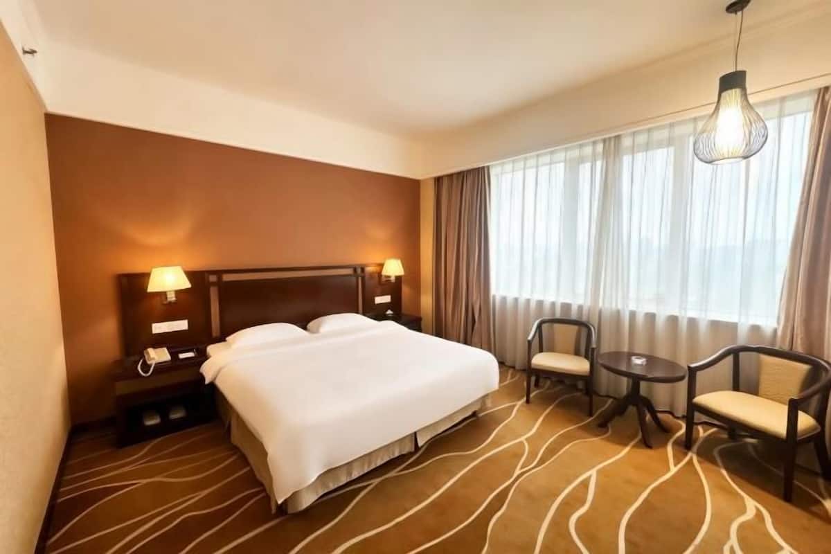 A hotel room with a large bed, two bedside tables with lamps, a small round table, two chairs, and a view of the outdoors through large windows.