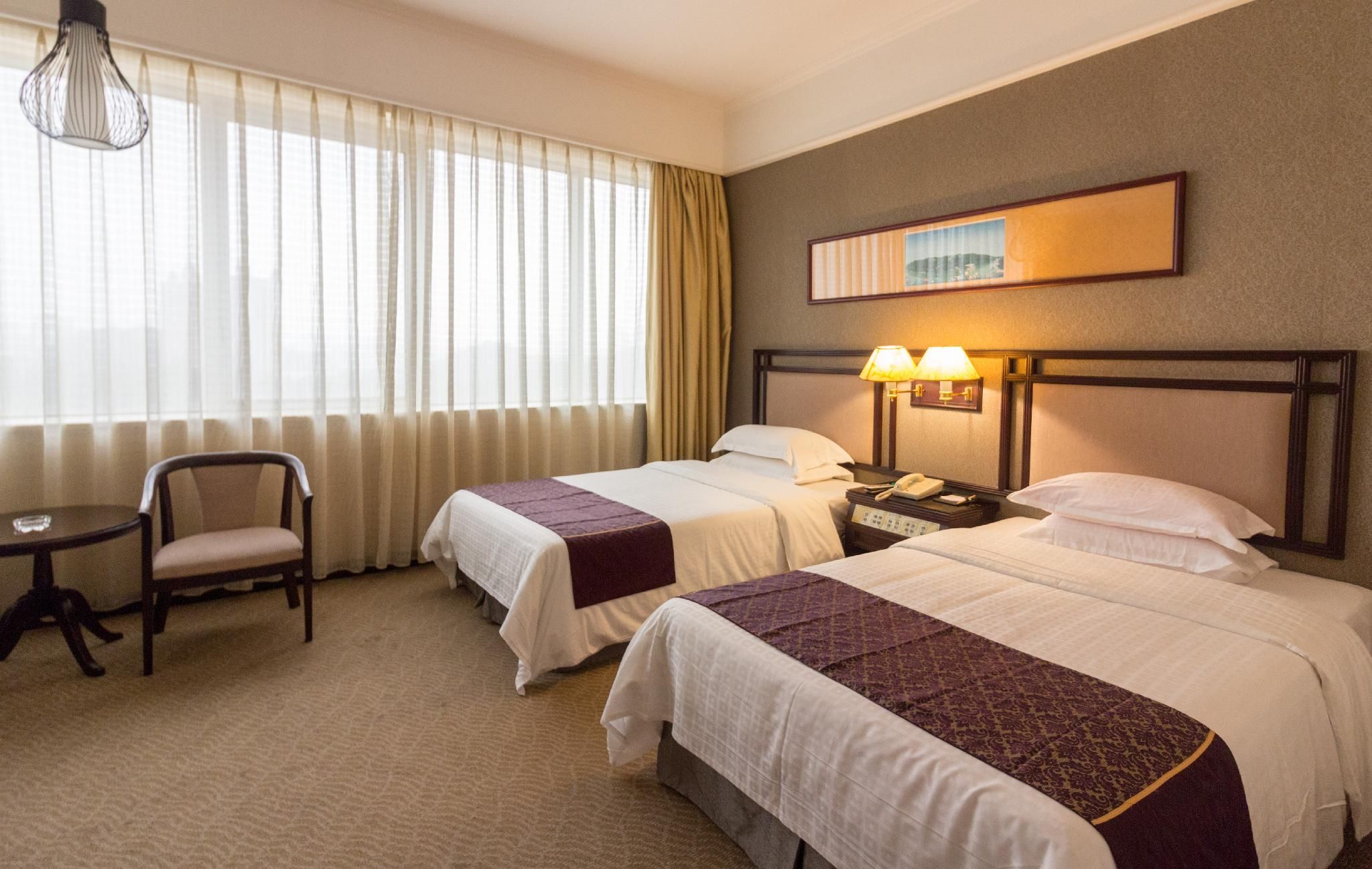 Una habitación de hotel con dos camas, una silla, una mesita pequeña y una mesita de noche con lámpara.