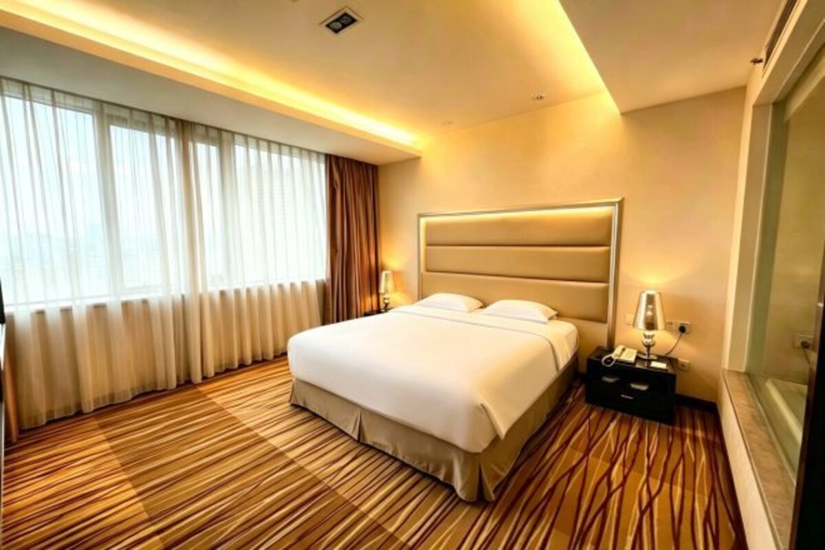 A hotel room with a large bed, a bedside table with a lamp, a window with curtains, and a wooden floor.