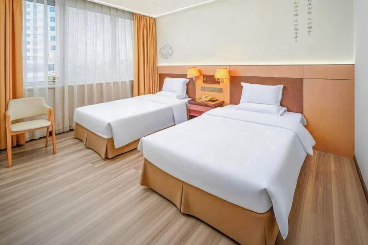 A hotel room with two beds, a chair, a nightstand with lamps, and a view of buildings through the window.