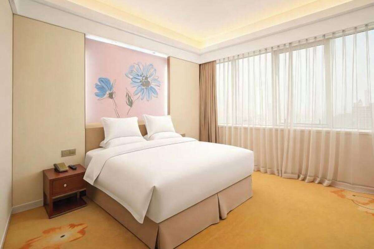 A hotel room with a bed, a nightstand, a painting on the wall, and a window with curtains.