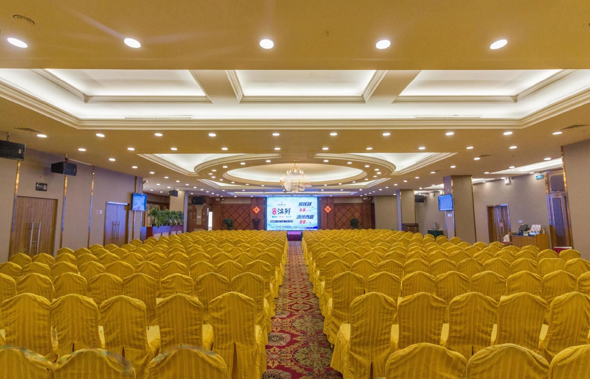A conference room with rows of yellow chairs, a stage with a screen, and a chandelier.