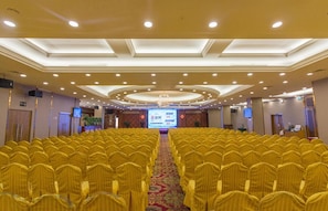 A conference room with rows of yellow chairs, a stage with a screen, and a chandelier. - Celeste Palace Hotel (Jiangmen)