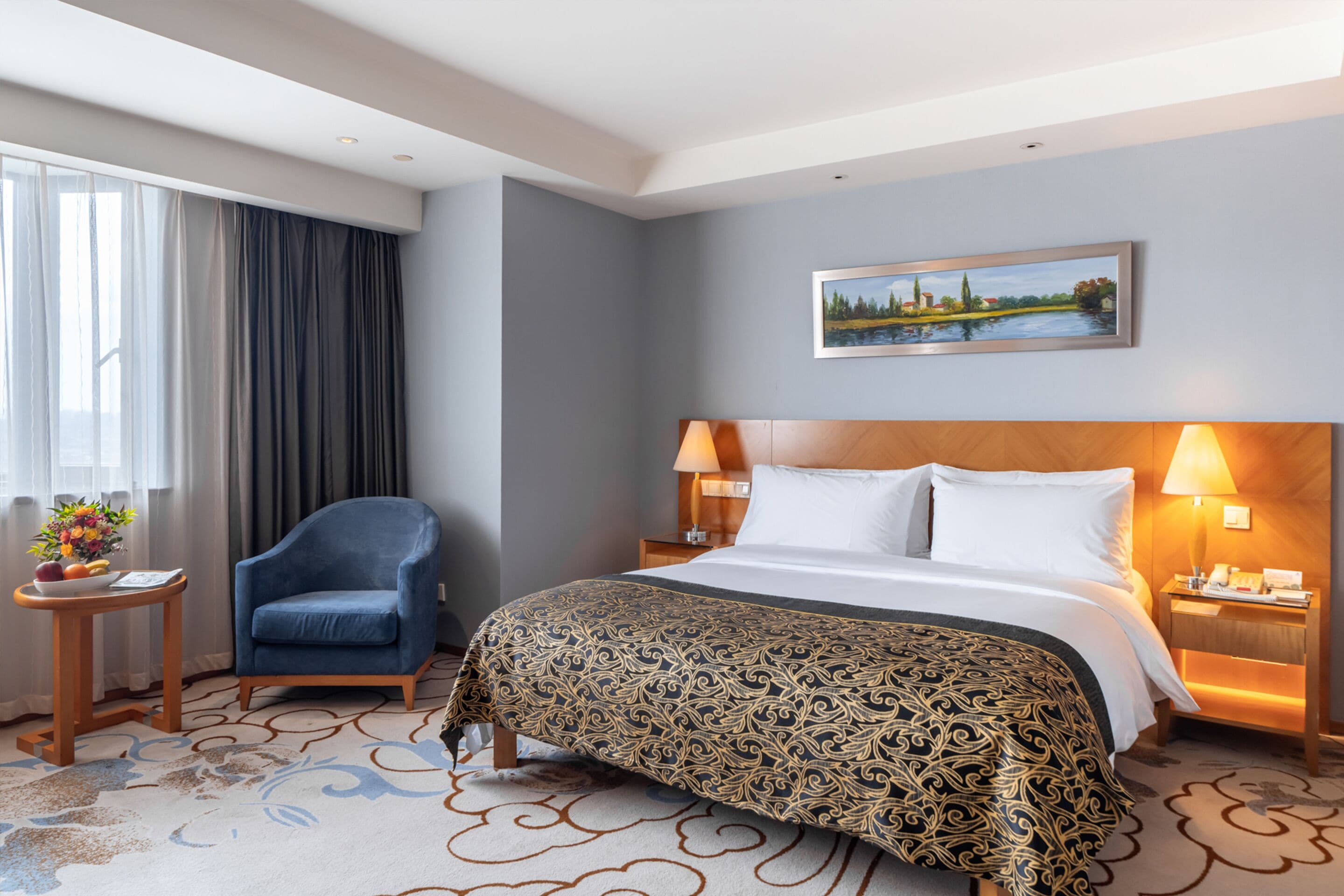 Una habitación de hotel con cama, una silla, una mesita de noche, una lámpara, un cuadro en la pared y una ventana con cortinas.