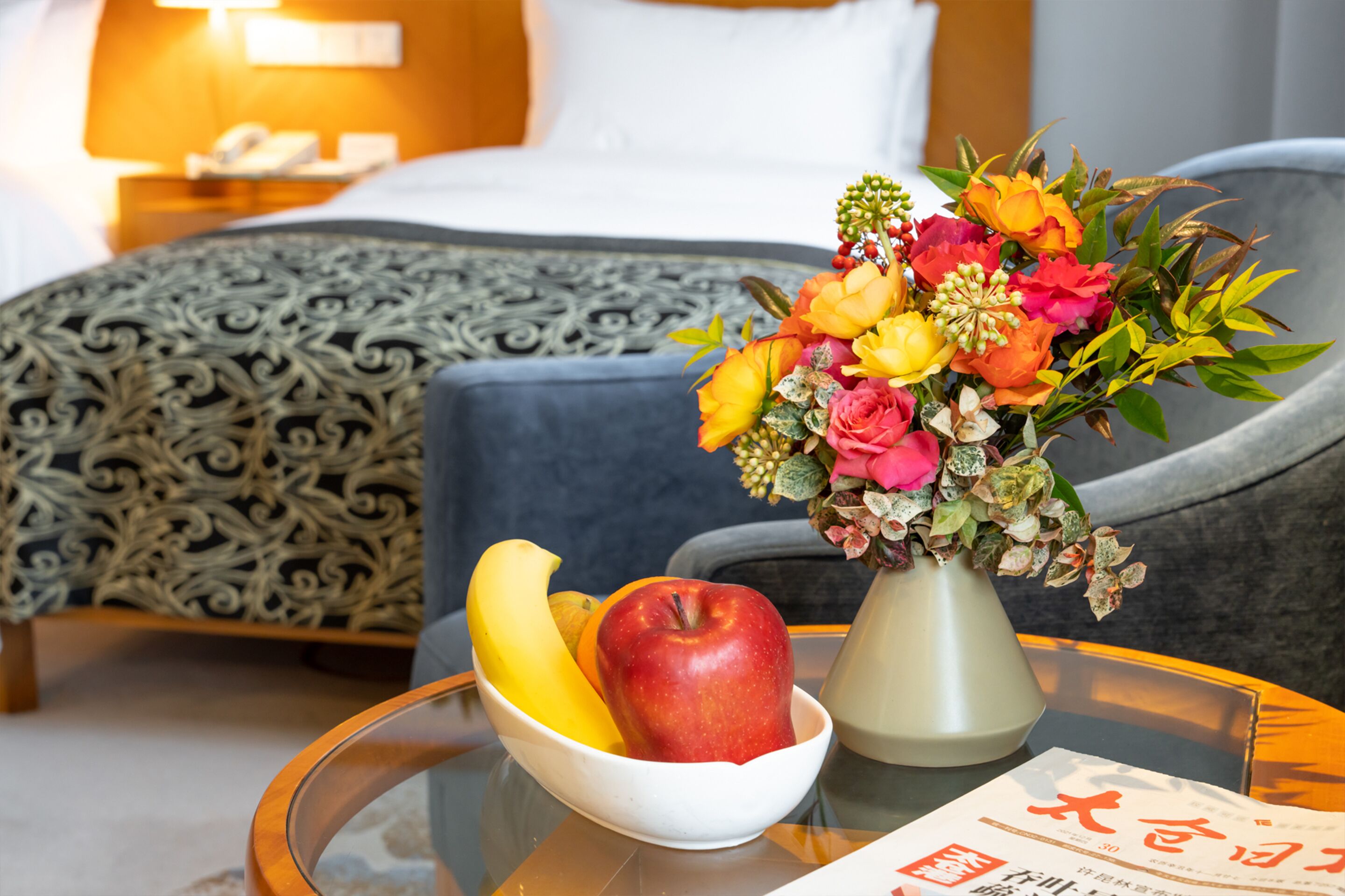 A hotel room with a bed, a bedside table with a bowl of fruit and a vase of flowers, and a magazine on the table.