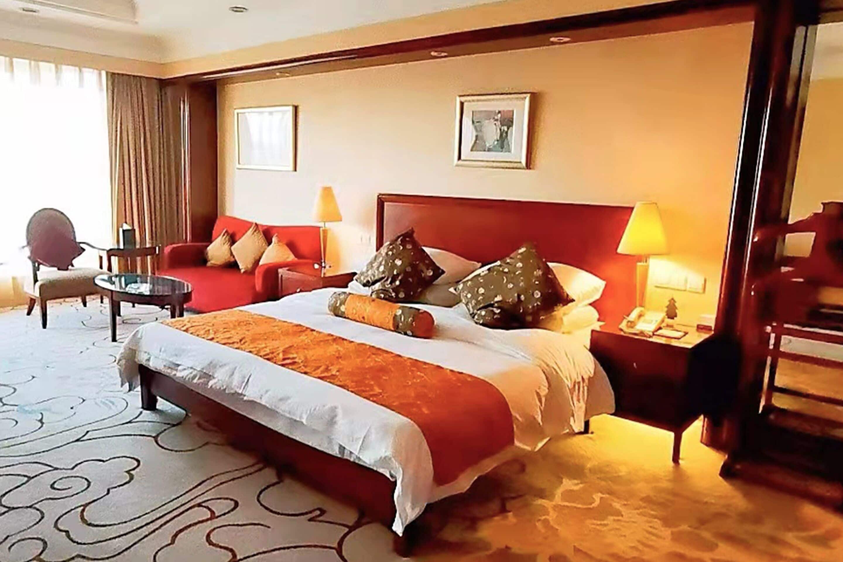 A hotel room with a large bed, a red sofa, a small round table, and a chair.