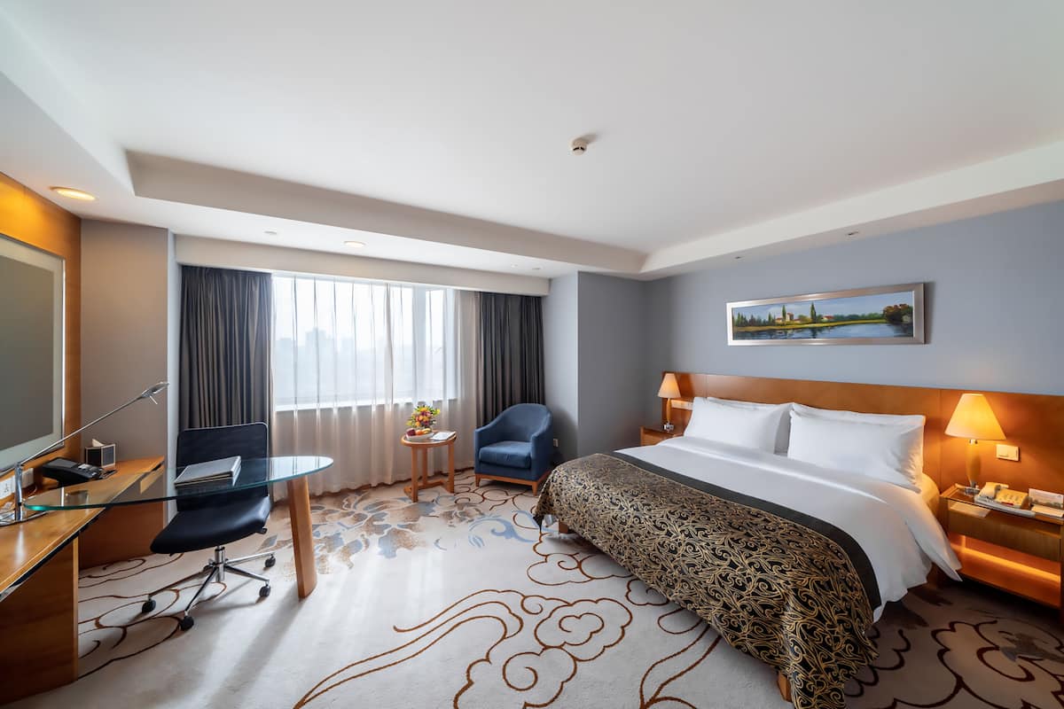 Une chambre d’hôtel avec un lit, un bureau avec un ordinateur, une chaise, une petite table avec une lampe et un tableau au mur.